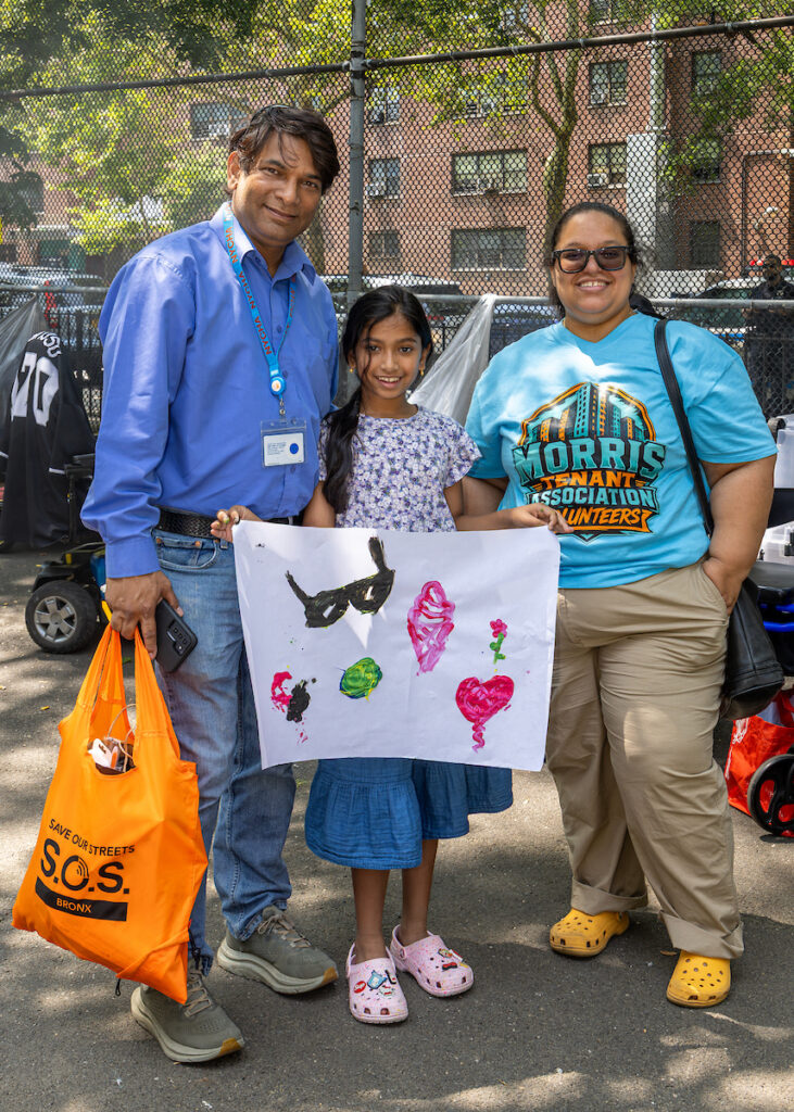 A family of three stands outdoors holding a child’s colorful artwork, with one adult carrying a tote bag and another wearing a volunteer shirt.