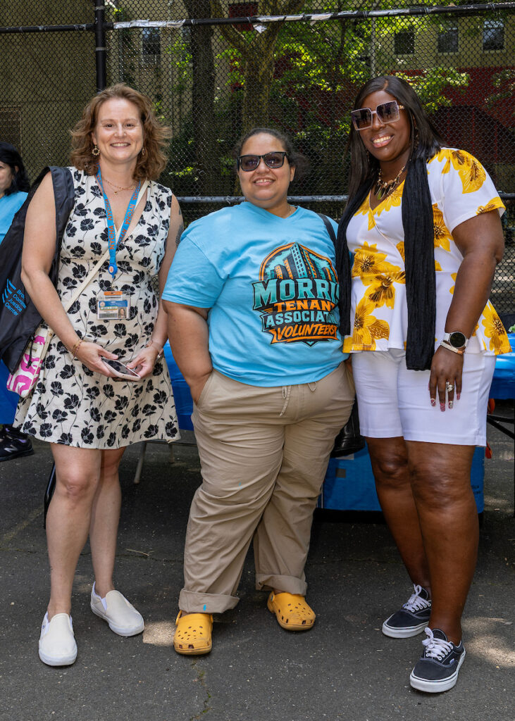 Three individuals pose together outdoors, one wearing a “Morris Tenant Association Volunteers” shirt, in front of a chain-link fence.