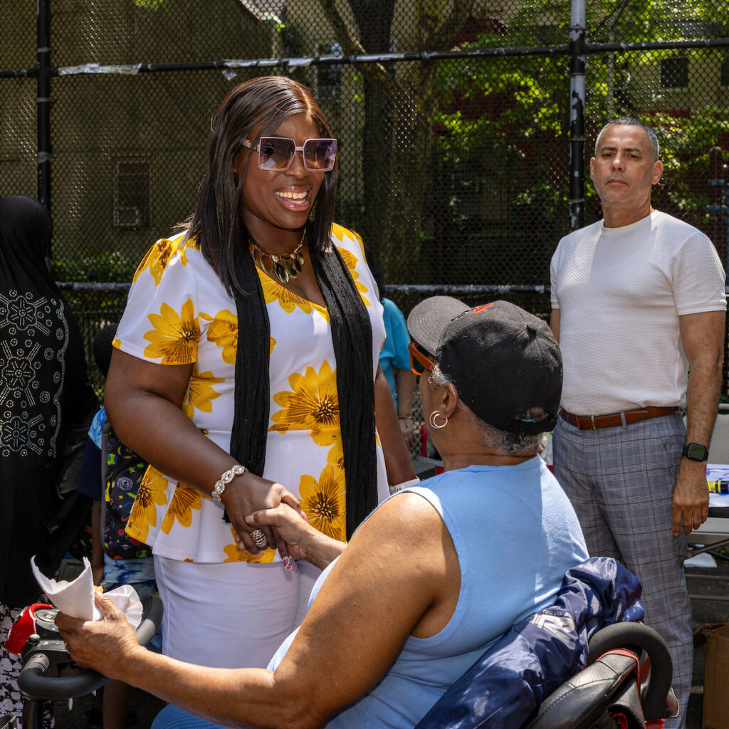 A person in a floral shirt and sunglasses smiles while shaking hands with someone in a wheelchair, with others standing nearby in an outdoor setting.