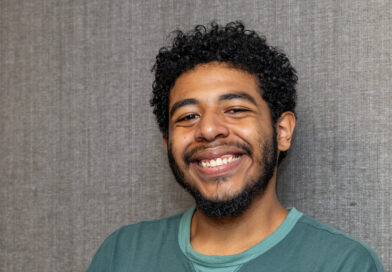 A person with curly hair and a beard smiles warmly while wearing a teal sweatshirt against a textured gray background.