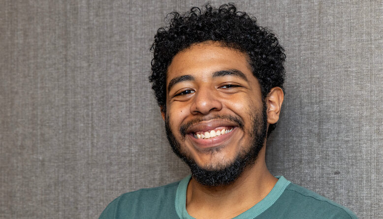 A person with curly hair and a beard smiles warmly while wearing a teal sweatshirt against a textured gray background.