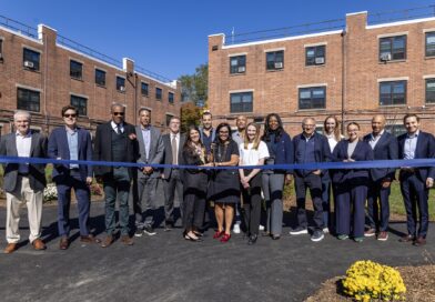 A group of people stand together outdoors, holding a blue ribbon during a ribbon-cutting ceremony in front of a brick building.