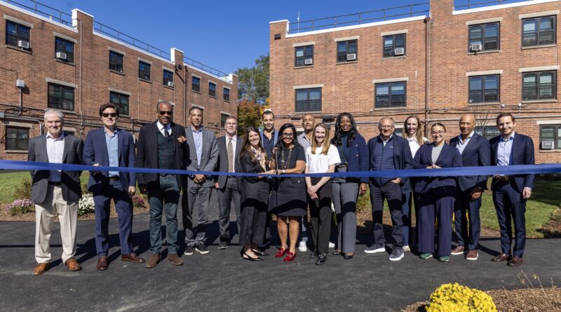 A group of people stand together outdoors, holding a blue ribbon during a ribbon-cutting ceremony in front of a brick building.