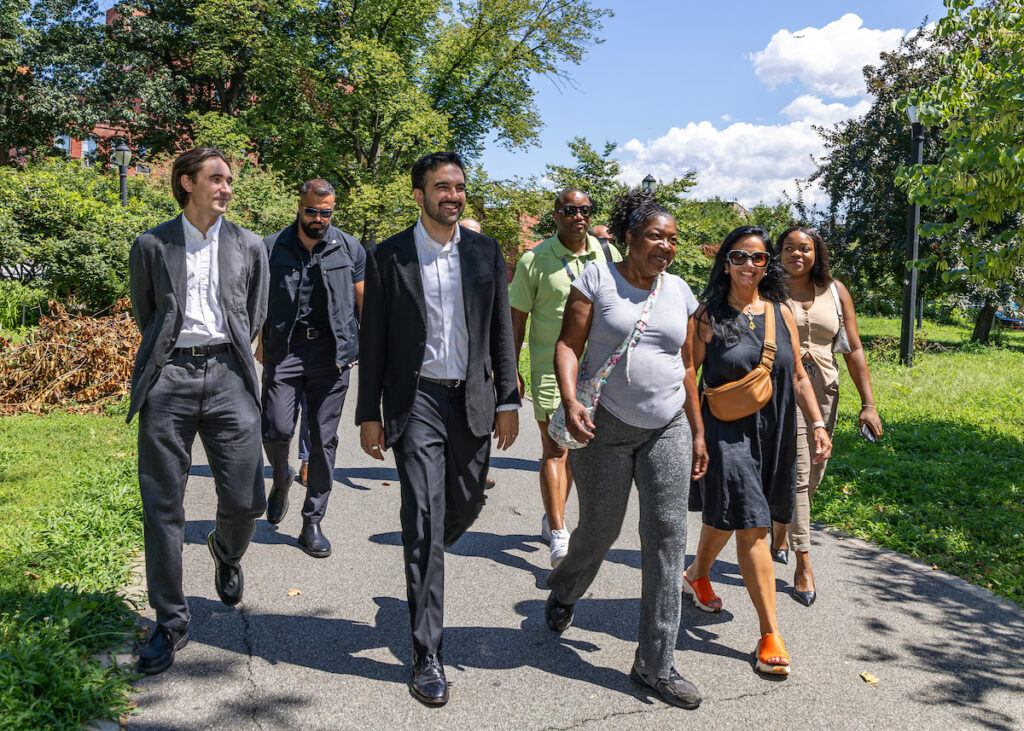 A group of people walk together along a paved path in a sunny park, surrounded by green trees and grass.