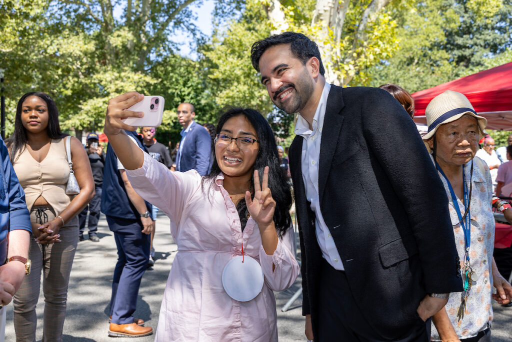 A young person takes a selfie with a smiling adult in a park, surrounded by other people and trees.
