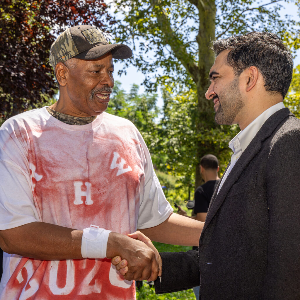 Two individuals shake hands outdoors, one wearing a camouflage cap and a red-stained shirt, the other in a dark suit, with trees and sunlight in the background.