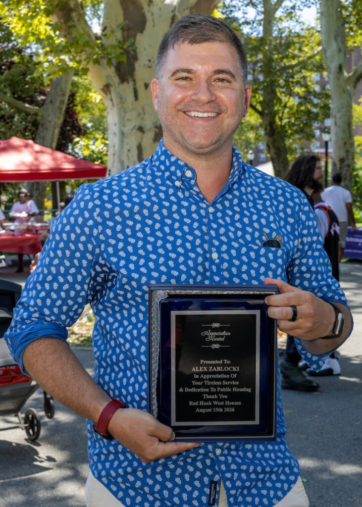 A person in a blue patterned shirt smiles while holding an “Appreciation Award” plaque presented to Alex Zablocki for their service to public housing.