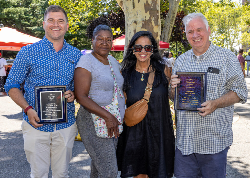 Four individuals stand outdoors, smiling and holding framed plaques, with trees and red tents visible in the background.