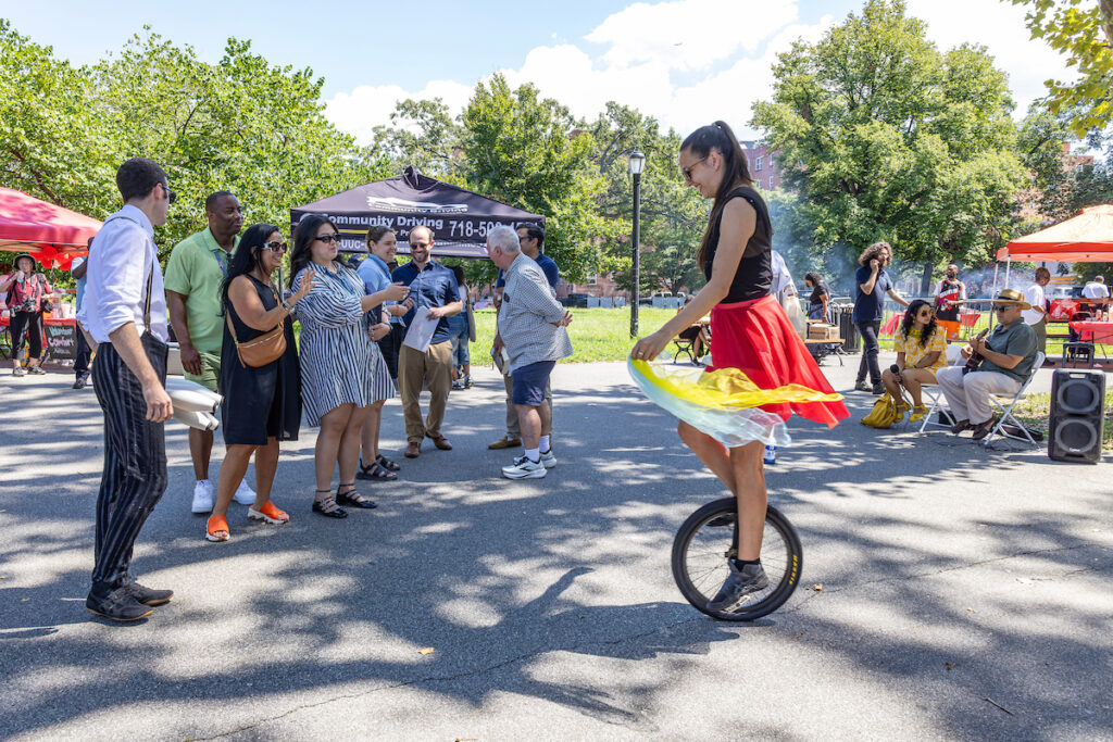 A person rides a unicycle in a park while a group of people watches, with trees, tents, and other attendees in the background.