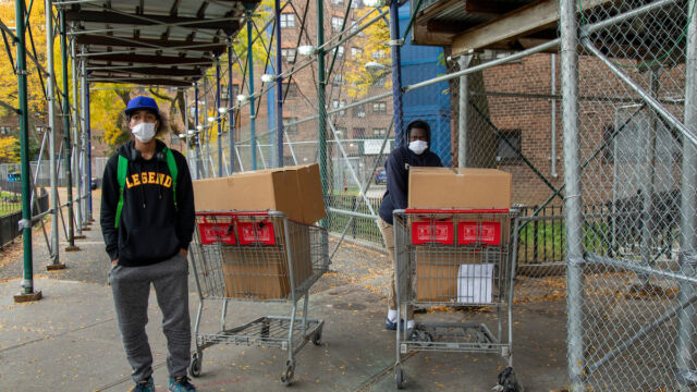 Two young residents from Astoria Houses volunteered to help distribute meals to residents. two men stand near shopping carts with cardboard boxes in them